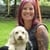 Woman with pink hair smiles while holding a small white dog in a grassy outdoor setting.